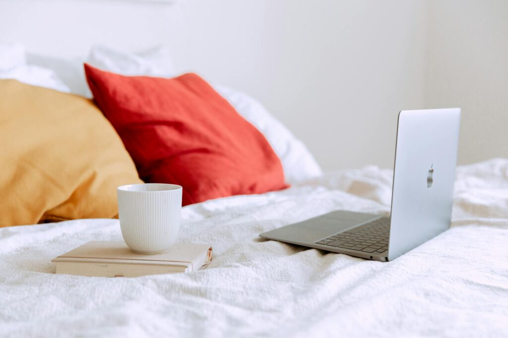 laptop and cup of coffee on notepad near pillows on bed at daytime