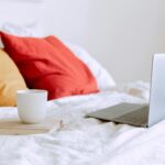 laptop and cup of coffee on notepad near pillows on bed at daytime